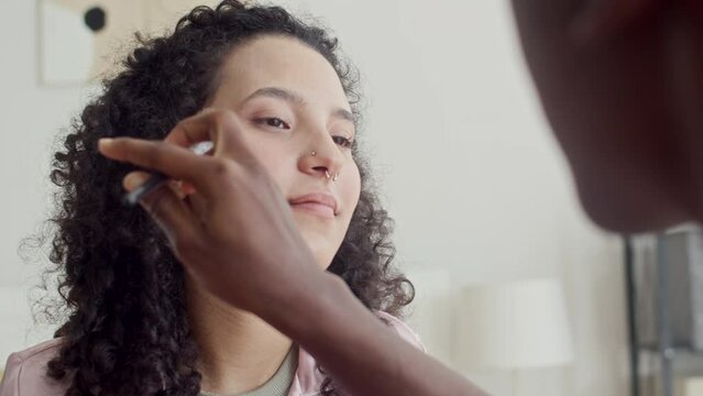 Over the shoulder of girl using brush when applying foundation on face of multi-ethnic girlfriend with piercing in nose