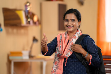 confident young Indian girl student with back pack showing thumbs up by looking camera at home - concept of education, aspirations and inspiration