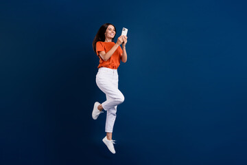 Full length photo of shiny cheerful lady wear orange t-shirt jumping chatting modern device empty space isolated dark blue color background