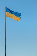 A large national flag of Ukraine flutters in the wind on a flagpole against the background of a clear blue sky