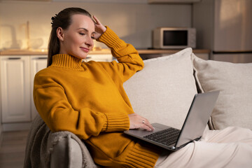 A contented woman dressed in a vibrant yellow sweater relaxes on a cozy sofa, deeply focused on her laptop screen in a warm, softly lit living area