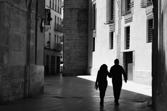 A Black and white photo with silhouette of a romantic couple walking on an urban European street in sunny daylight. 