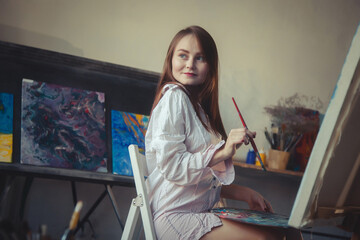 Smiling woman painter with brush and palette sitting at canvas, drawing looking away. Happy lady artist in white shirt painting oil paints in art lab. Artisanal lifestyle concept. Copy ad text space