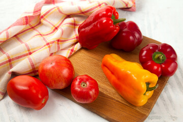 Cutting board with several red and yellow tomatoes and bell pepper on white wooden background with red kitchen towel..