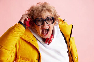 Emotional senior woman listening music in headphones and singing wearing yellow puffer jacket....