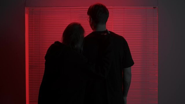 Portrait of male model in dark studio. Young man in t-shirt standing near window with red light behind jalousie, girl comes and hugs man.