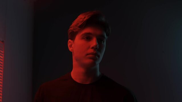 Portrait of male model in dark studio. Young man in t-shirt standing near window with red light behind jalousie looks sideway from camera.