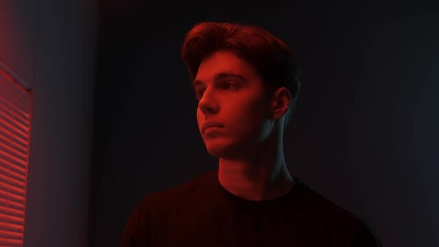 Portrait of male model in dark studio. Young man in t-shirt standing near window with red light behind jalousie looks at sides from camera.