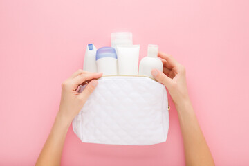 Young adult woman hands holding white cosmetic travel bag with plastic bottles on pink table...