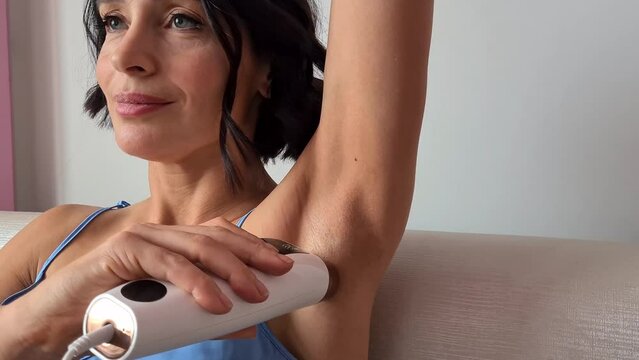 Young woman epilating armpits with modern photoepilator in bedroom at home concept