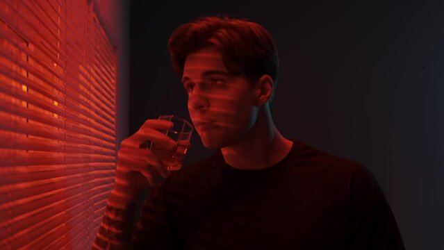 Portrait of male model in dark studio. Young man in t-shirt standing near window with red light behind jalousie drinks beverage opens blinds.