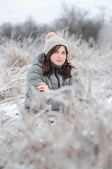 Winter portrait of young woman baby face in winter clothes sitting on field on frosty day. Pretty girl on nature