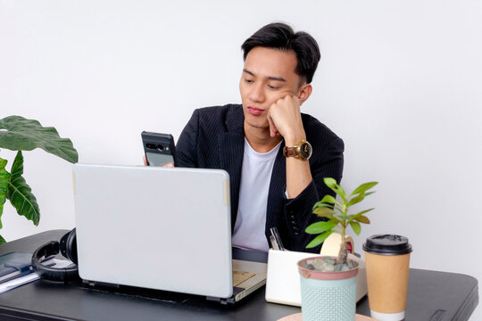 A young Asian man appears bored while multitasking with his smartphone and laptop at a modern workspace. Dispirited and lacking motivation.