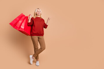 Full length photo of funky dreamy lady dressed red pullover holding shoppers pointing empty space isolated beige color background