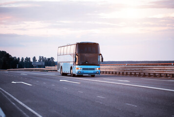 A blue comfortable bus carries passengers along the highway in the evening with the headlights on....