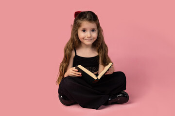 Portrait of a smiling little girl sitting with legs crossed, studying isolated over pink background