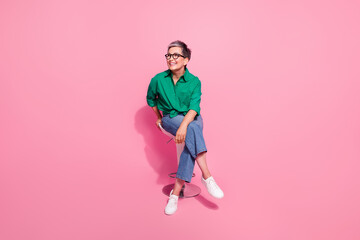 Full body photo of pleasant nice woman wear green shirt look at proposition empty space sit on stool isolated on pink color background - obrazy, fototapety, plakaty