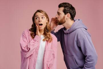 Naklejka premium Shock, gossip, share advice. Young man whispering to lady on ear, happy woman with open mouth excited and surprised, posing on pink studio background 