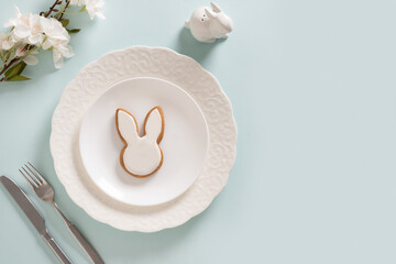 Easter table setting with white plate, glazed cookies and white bunny on blue background. View from above. Space for greeting text.