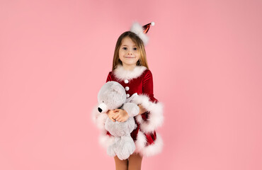 Lovely little girl in Christmas dress isolated on pink background holds her teddy bear in her arms and smiles beautifully.