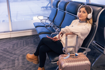 Caucasian young woman sitting at the airport terminal while waiting for boarding for vacation. Uses smartphone to listen music with headphones while looks at the camera with a carry-on suitcase 
