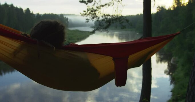 Man with dreadlocks from back swinging in a colorful hammock watching a river bend during dusk. Shot on RED.