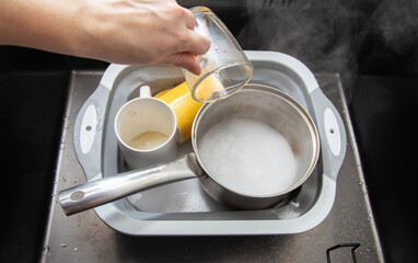 A woman's hand pours hot water into a basin with dirty dishes and environmentally friendly oxygen bleach. Washing dishes from grease and dirt. - obrazy, fototapety, plakaty