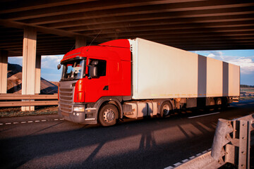 A red tractor with an isothermal refrigerated semi-trailer transports perishable goods with temperature conditions against the backdrop of sunset under the bridge. Delivery of perishable products