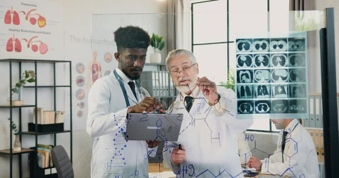 Doctor which standing near glass wall with attentive smart man worker and discussing depicted chemical formula