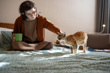 Girl petting Devon Rex cat sits in bed at home drinking coffee at home. Woman stroking domestic...