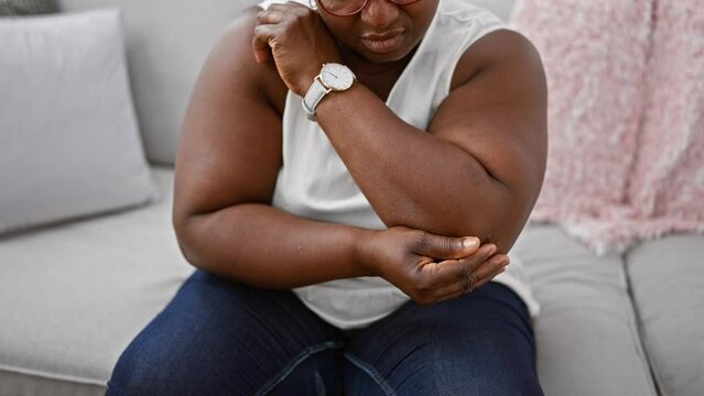 Unhappy african american woman, relaxing on comfy sofa at home, bravely bearing pain from elbow injury. her worried expression shines through cool glasses.