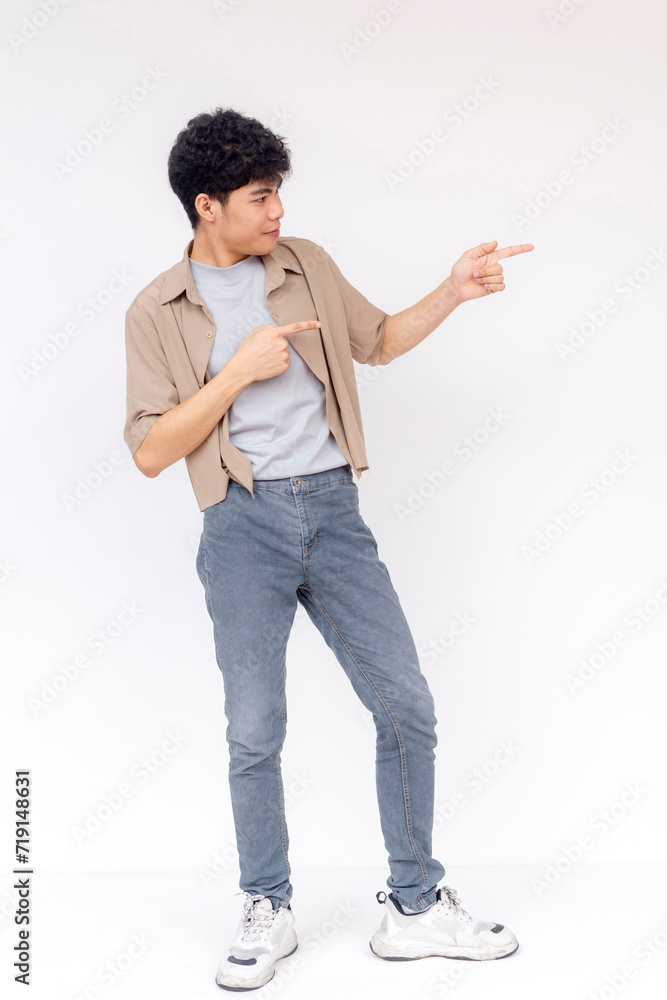 Wall mural A happy young Asian man with curly hair pointing to the side, smiling and standing against a white studio background.