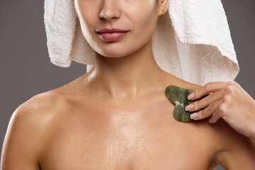 Beautiful young woman in towel on head, after shower doing body massage with gua sha against studio...