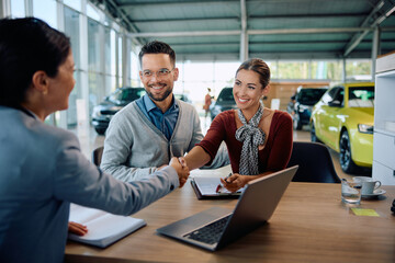 Happy couple shaking hands with saleswoman after buying new car in showroom.