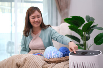 Close up at part of air purifier and Asian woman hand press touch open button while knitting on sofa at home with smile of happiness and appreciate felling.