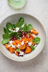 Plate with roasted sweet potato, fresh spinach, feta and pomegranate salad, vertical shot on a beige stone background, view from above