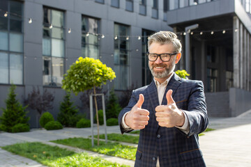 Smiling senior professional in a suit giving thumbs up outside an office building, representing...