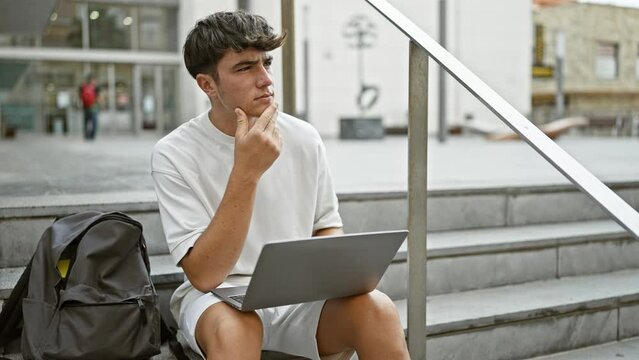 Deep in thought, a young, handsome hispanic teenager student, absorbed in his laptop, sitting casually on city university stairs in relaxed concentration