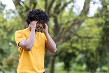 Close-up photo of a young Indian sportsman standing in the park and holding his head with his...