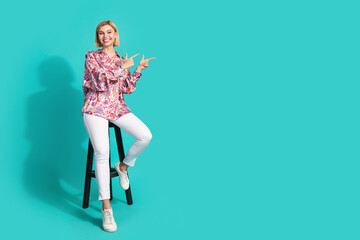 Full length photo of nice young lady point empty space sit chair wear trendy pink print garment isolated on cyan color background