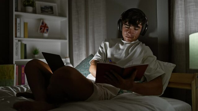 Confident young hispanic teenager, diligently studying online via laptop, taking notes while sitting on bed, lighting up his bedroom with a radiant smile during a late-night video call