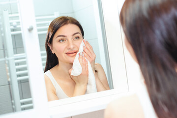 Beautiful woman in a white tank top wiping her face with a white towel while looking in the mirror...