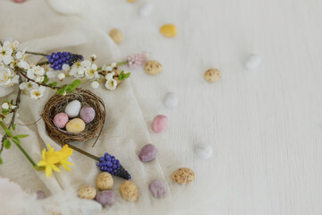 Obraz na płótnie Canvas Colorful easter chocolate eggs in nest, spring flowers, feathers and linen border composition on white wooden table. Space for text. Happy Easter! Seasons greetings