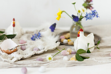Happy Easter! Stylish easter chocolate eggs, spring flowers, chicken figurines and linen cloth on rustic wooden table. Space for text. Easter modern simple decoration