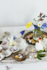 Fototapeta na wymiar Colorful easter chocolate eggs in nest, spring flowers, chicken figurine and linen cloth on rustic wooden table. Space for text. Easter modern simple decoration. Happy Easter!