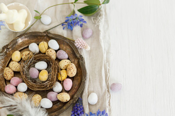 Fototapeta na wymiar Happy Easter! Colorful easter chocolate eggs in nest, spring flowers, feathers and linen cloth on rustic wooden table. Space for text. Easter modern simple decoration