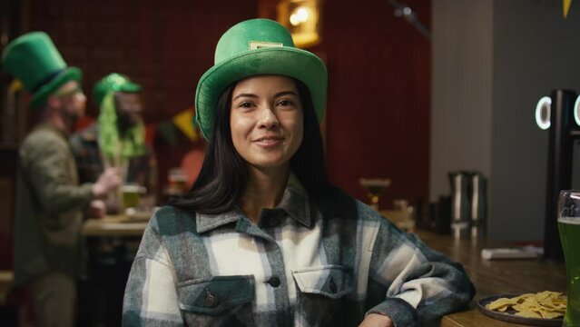 Chest up portrait of pretty smiling Biracial woman wearing green leprechauns hat and posing for camera near counter with green beer in Irish pub