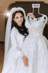A beautiful bride with a long veil in her room, wearing a robe. Wedding dress on a mannequin. The...