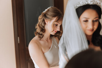 Fototapeta premium Wedding morning. Bridesmaids help put on the white wedding dress. A young woman is preparing to meet her groom and having fun with her friends