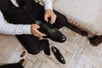 Fototapeta premium A young man put on black leather boots indoors. Close-up photo. Detail of a groom putting on his shoes